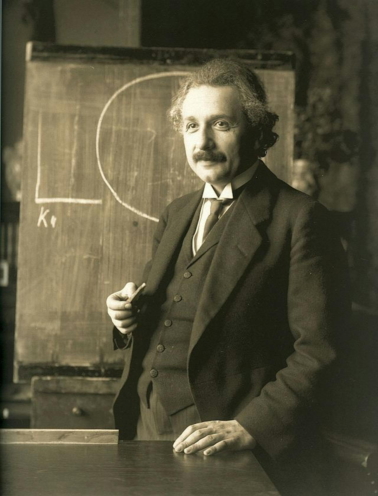 Albert Einstein staat voor een schoolbord. De foto is in sepia.