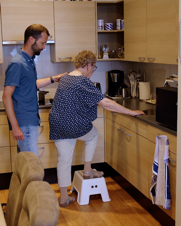Ergotherapeut helpt oudere vrouw op een opstapje in de keuken.