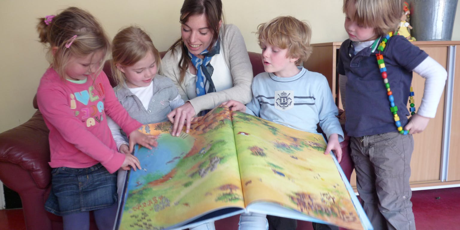 Een volwassene zit met een groep kinderen op de bank een boek voor te lezen.