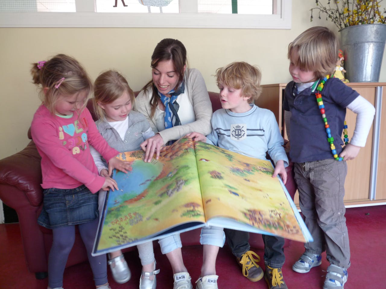 Een volwassene zit met een groep kinderen op de bank een boek voor te lezen.