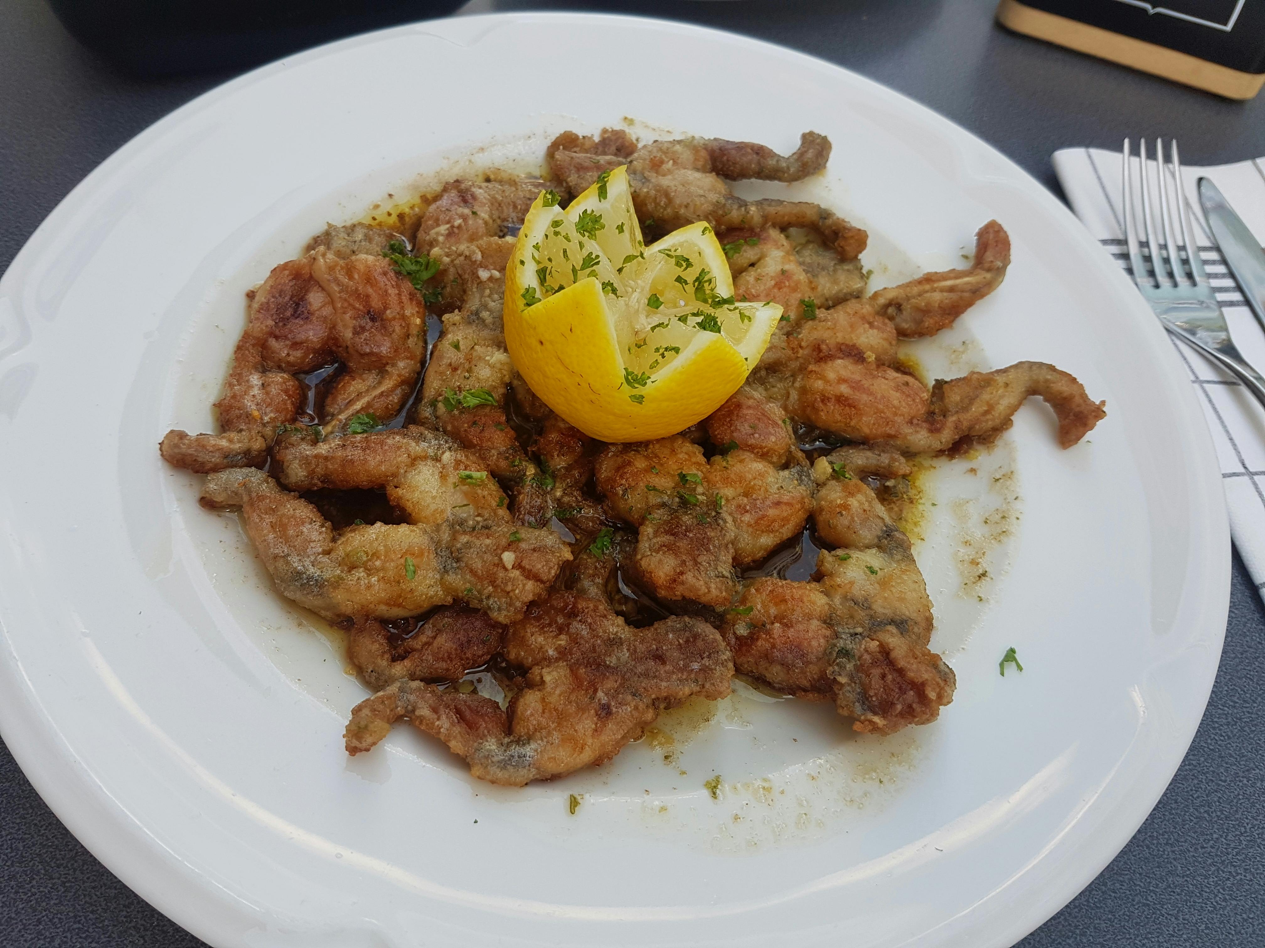 Een wit bord met daarop een berg gepaneerde, gebakken kikkerbillen. Bovenop de kikkerbillen ligt een citroen die gesneden is in de vorm van een kroon.