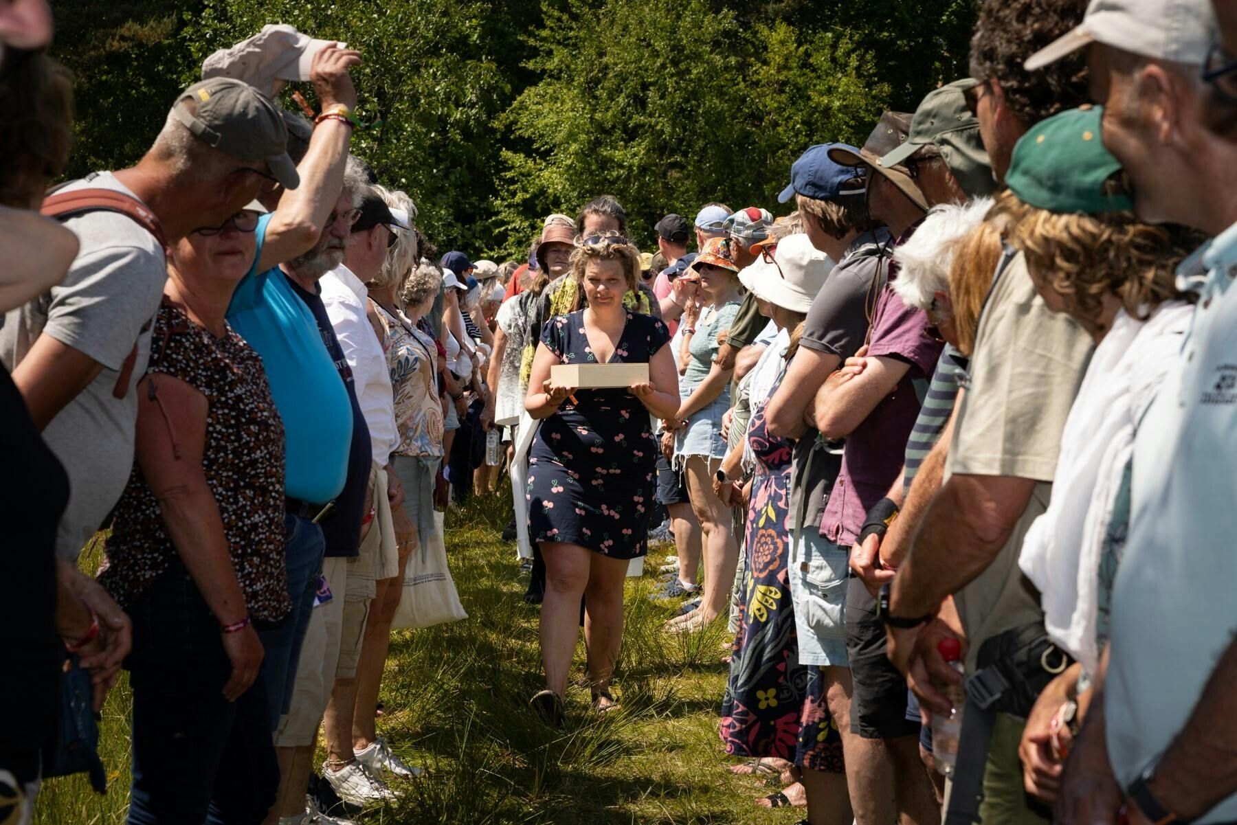 Een erehaag van mensen staat in een grasveld. In de verte loopt een jongedame in een zomerse jurk richting de camera. Ze draagt een houten doos in haar handen.