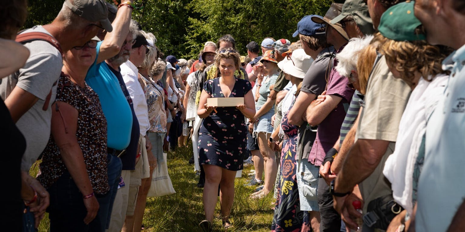 Een erehaag van mensen staat in een grasveld. In de verte loopt een jongedame in een zomerse jurk richting de camera. Ze draagt een houten doos in haar handen.