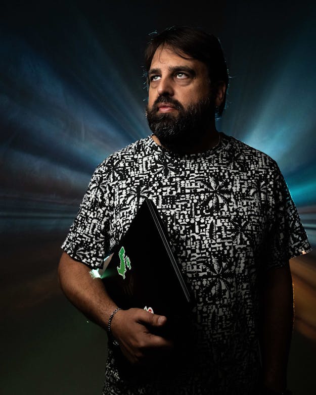 Een man met baard heeft een laptop onder zijn arm. De achtergrond is zwart. Van achter zijn rug komen lichtstralen tevoorschijn. Een man met baard heeft een laptop onder zijn arm. De achtergrond is zwart. Van achter zijn rug komen lichtstralen tevoorschijn.