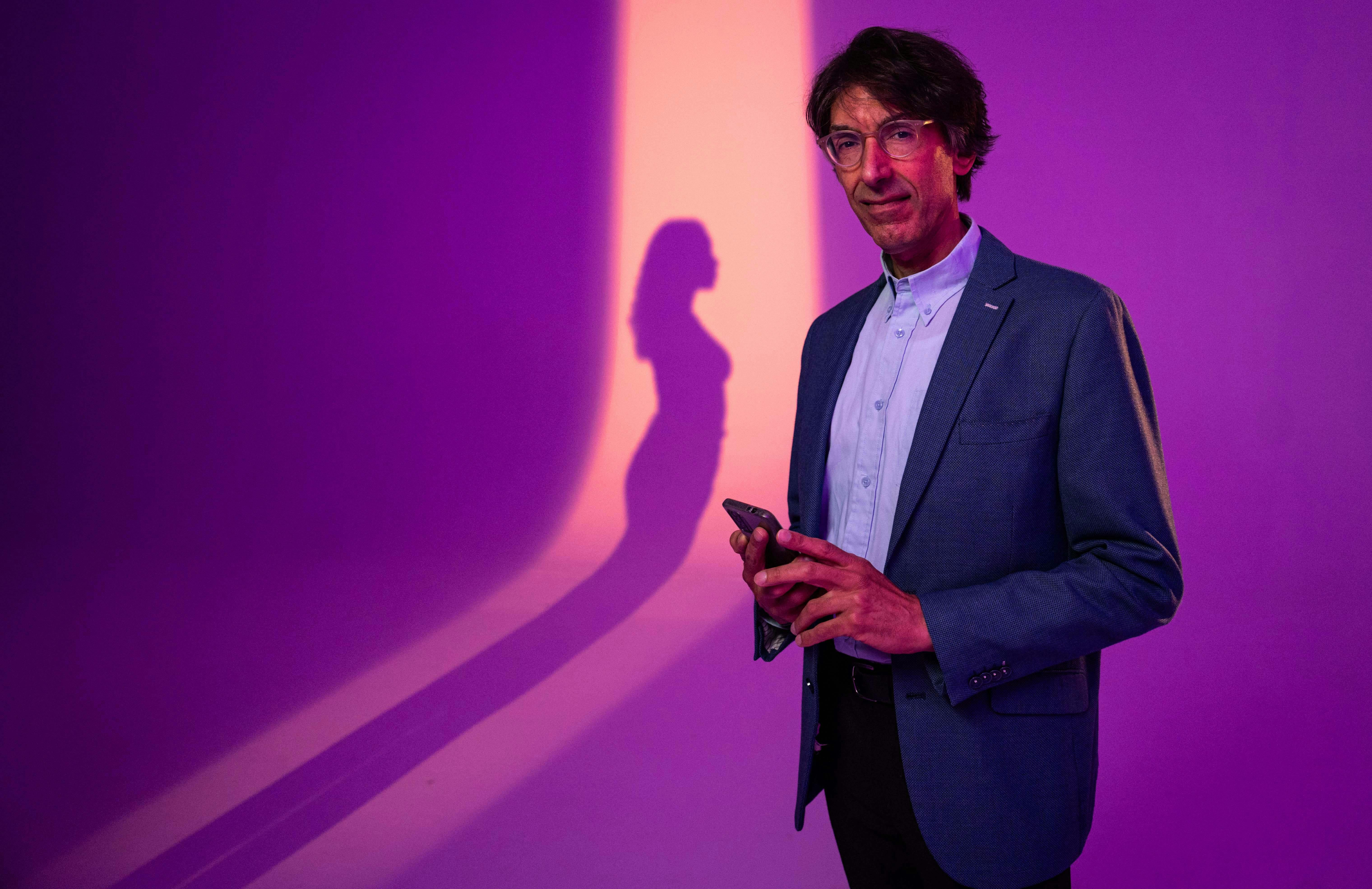 Een man staat met een telefoon in zijn hand. Op de achtergrond is de schaduw van een vrouw te zien.