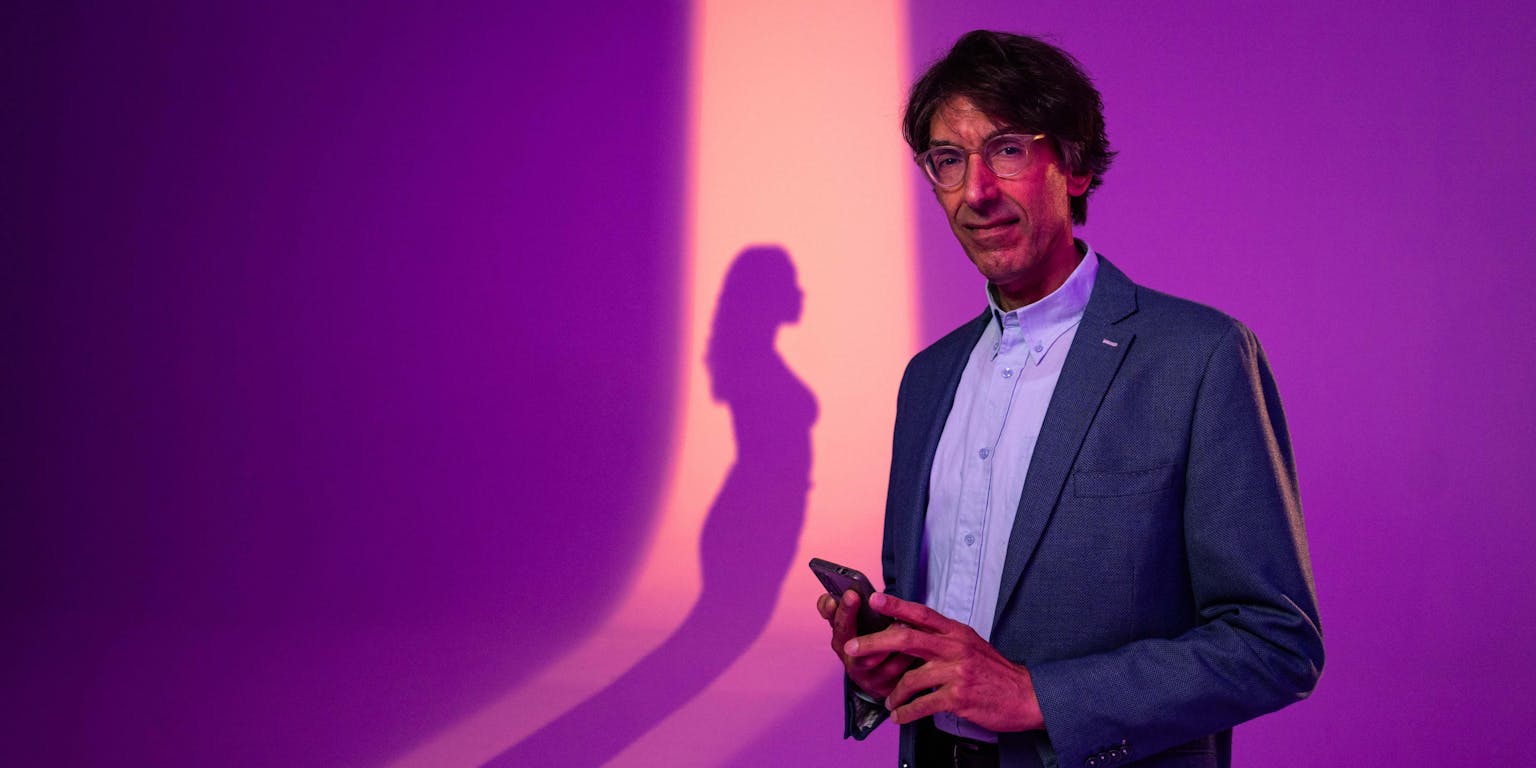 Een man staat met een telefoon in zijn hand. Op de achtergrond is de schaduw van een vrouw te zien.