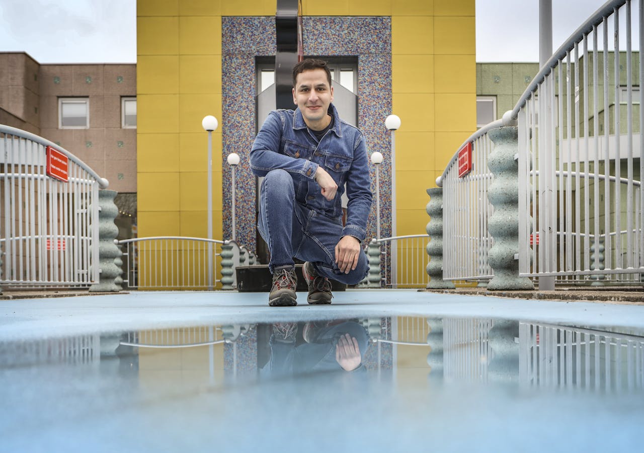 Een man in spijkerkleding zit buiten op een brug op zijn hurken. Links en rechts is een lichtgrijs hekwerk te zien. OP de achtergrond een geel met grijs gebouw.