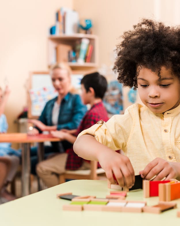 Hoogbegaafd potentieel is een combinatie van hoge intellectuele capaciteiten, een groot creativiteit denkvermogen en een grote (leer)motivatie Kleuter met donkere krullen speelt met blokjes met op de achtergrond een juf met drie andere kinderen.