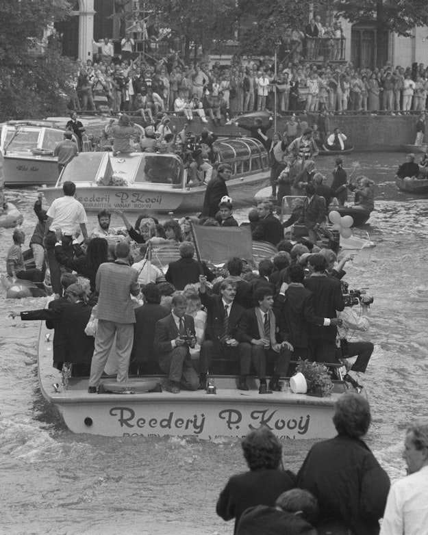 Een rondvaartboot met daarop het Nederlands elftal vaart door de Amsterdamse grachten. Op de kades staat veel publiek de voetballers toe te juichen. Op het water varen ook nog andere boten vol mensen. Het is een zwart-witfoto.