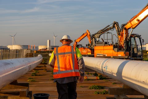 Werkzaamheden aan het landelijke waterstofnetwerk van Gasunie in de Rotterdamse Dintelhaven. Een man met bouwhelm en oranje veiligheidshesje loopt tussen twee grote witte pijpleidingen. Rechts is oranje graafmachine te zien.
