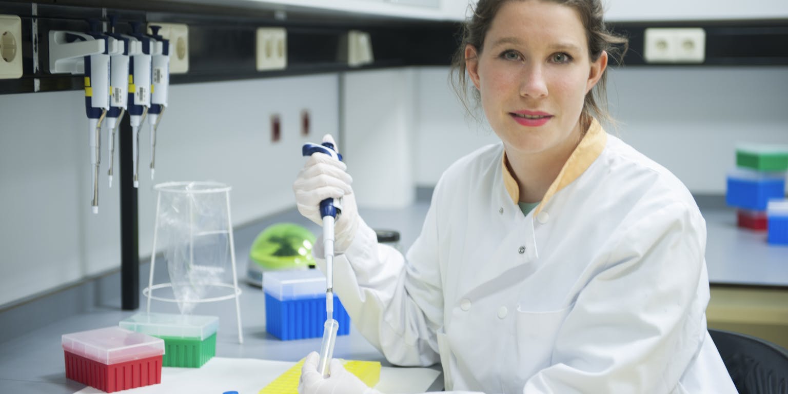 Karin, gekleed in een witte laboratoriumjas, gebruikt een pipetteerder om zorgvuldig een heldere vloeistof over te brengen in een reageerbuisje.