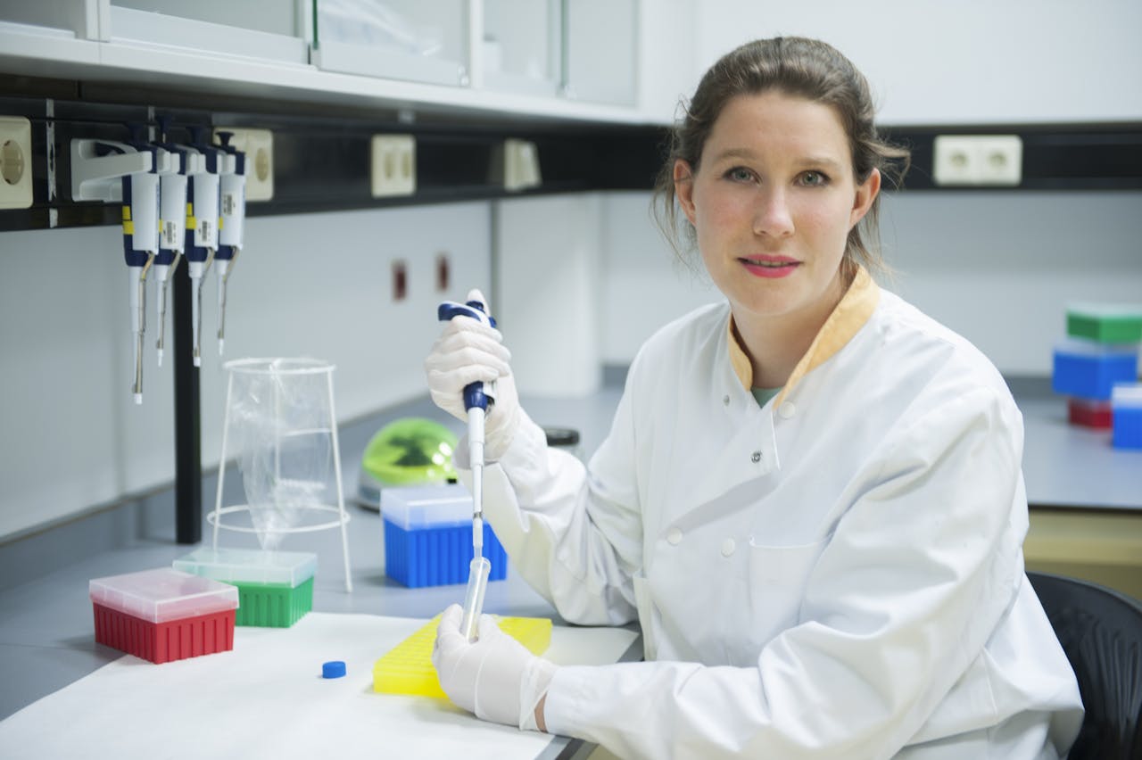 Karin, gekleed in een witte laboratoriumjas, gebruikt een pipetteerder om zorgvuldig een heldere vloeistof over te brengen in een reageerbuisje.