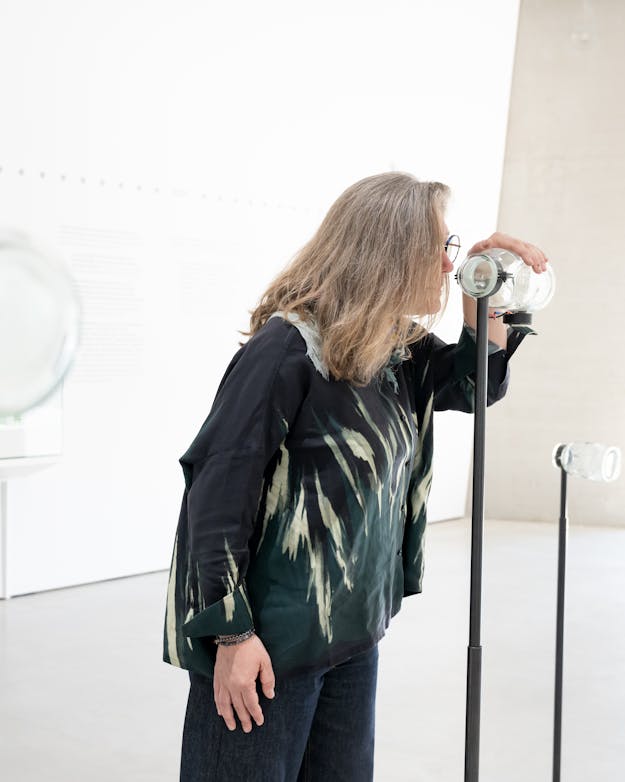 Een mevrouw ruikt aan een glazen pot op een standaard. Een mevrouw ruikt aan een glazen pot op een standaard.