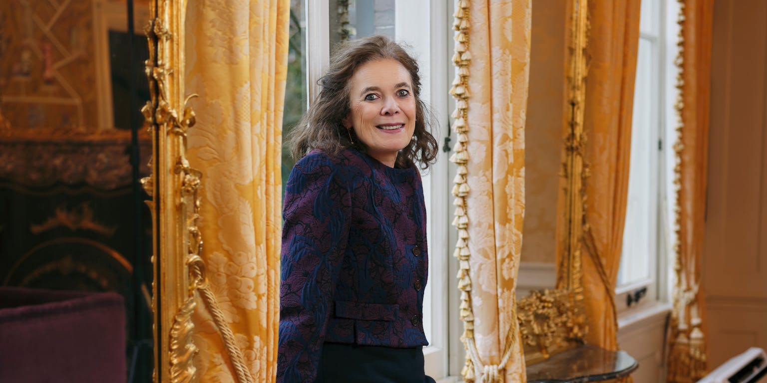Schrijfster Louise Fresco, een oudere blanke vrouw met donker haar, lacht naar de camera. Ze leunt tegen het raamkozijn van een rijkelijk versierde kamer met gouden lijsten en ornamenten.