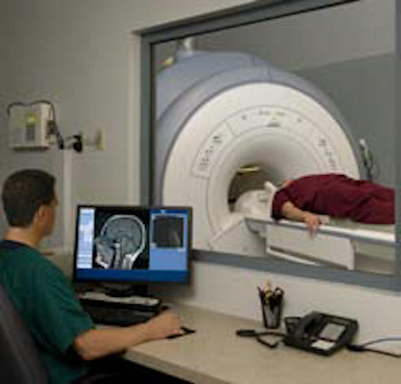 Een persoon ligt voor de MRI-scan. Een persoon zit aan de andere kant van het glas achter zijn bureau met een computerscherm voor zich.