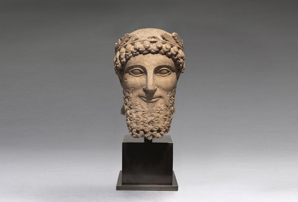 <p>Mannenportret, kalksteen, vijfde eeuw v.Chr., gevonden op Cyprus, h. 34 cm. Dit portret maakte ooit deel uit van een levensgroot beeld. Waarschijnlijk stond het beeld opgesteld bij een tempel of heiligdom, als geschenk voor de godheid die daar werd aanbeden. Vanwege de grote afmetingen was het kostbaar. De schenker moet behoord hebben tot een van de rijkste families van het vijfde-eeuwse Cyprus. In die periode stond het eiland onder bewind van de Perzen, die naast hun gezag ook hun beeld- en vormentaal meenamen naar de Griekse wereld. Dat laat zich zien in dit portret. Naast de typische Grieks-Archaïsche stijlkenmerken zoals de amandelvormige ogen en ‘archaïsche’ glimlach, heeft de man, geheel volgens de mode aan het Perzische hof uit die tijd, een gestileerde golvende baard en kapsel.</p>