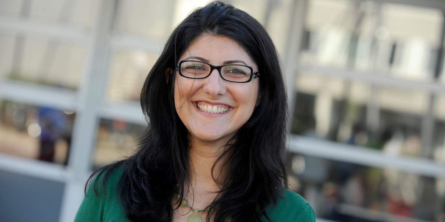 Een vrouw met een bril glimlacht voor een gebouw.