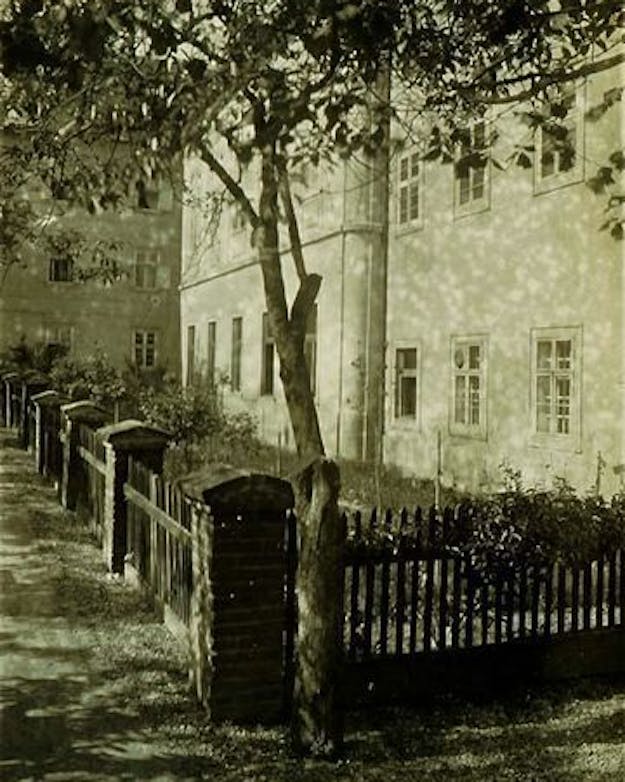 Mendel’s moestuin. Sepia-gekleurde foto van een omheinde tuin met een boom voor een wit gebouw.