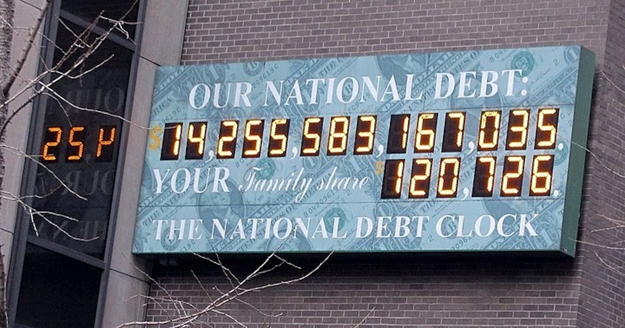 Aan een muur van grijze baksteen hangt een groenig bord met in witte letters 'Our national debt' en 'The National Debt Clock'. Digitaal worden cijfers weergegeven.
