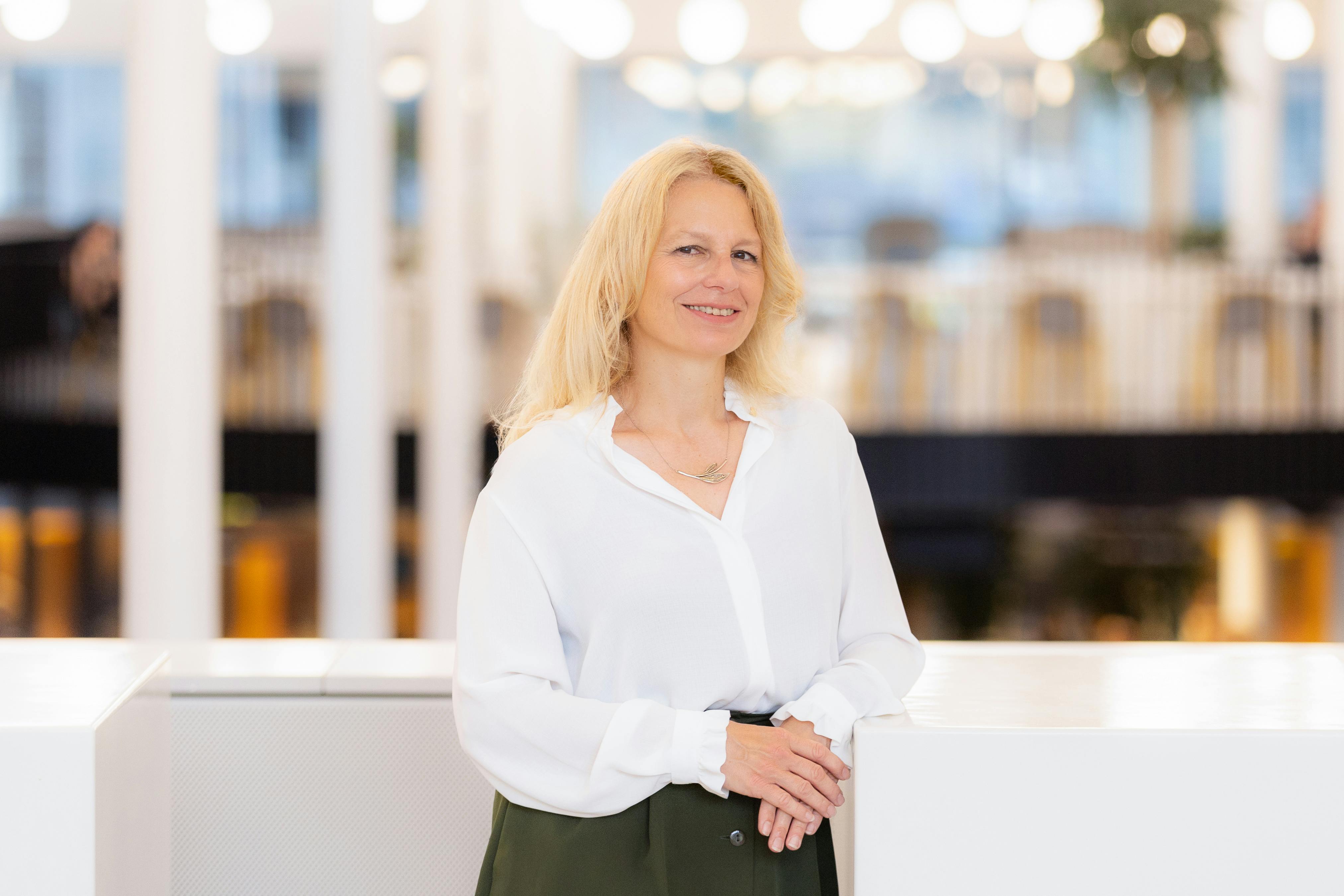 Portretfoto van Natali Helberger. Ze leunt op een witte balustrade. Op de achtergrond is vaag de binnenkant van een gebouw te zien met witte pilaren.