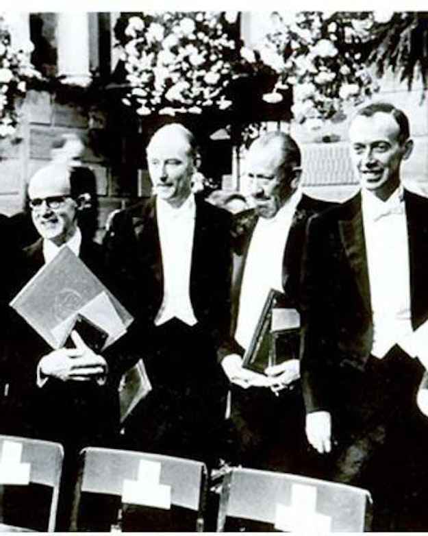 Zes Nobelprijswinnaars in gala-outfit naast elkaar lachend naar de camera voor een zwart-wit foto.