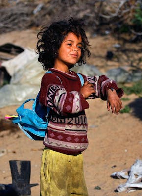 Een kind met vieze kleding aan met een blauwe rugzak op haar rug. Op de achtergrond is veel afval te zien.