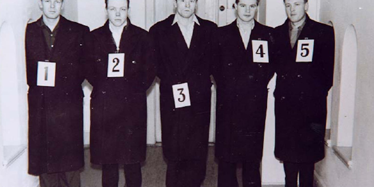 Een groep mannen in het zwart gekleed die naast elkaar staan. Om de nekken van de mannen hangen de cijfers 1 tot en met 5.