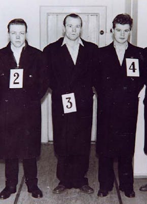 Een groep mannen in het zwart gekleed die naast elkaar staan. Om de nekken van de mannen hangen de cijfers 1 tot en met 5.