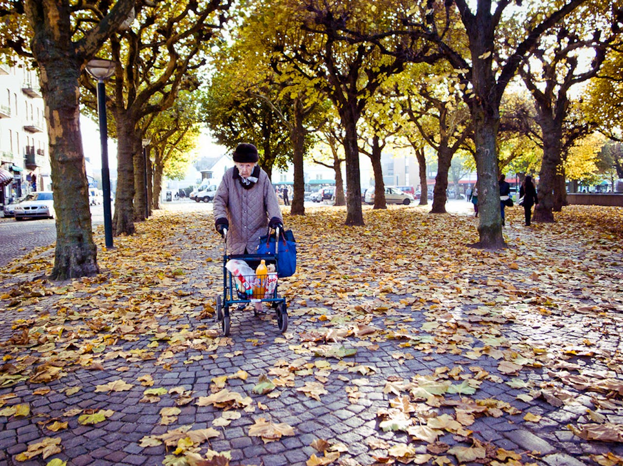 Een oude dame loopt door de straat met haar rollator. Er zijn veel bomen en er liggen bladeren op de grond.
