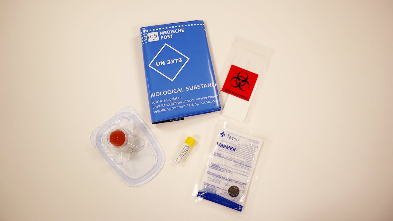 De verschillende onderdelen die je ontvangt in een PFAS zelftest, waaronder een bloedafname-apparaat, het buisje om het bloed in op te vangen en een retourenvelop. Foto van de onderdelen die je ontvangt in een PFAS zelftest, waaronder een bloedafname-apparaat, het buisje om het bloed in op te vangen en een retourenvelop.