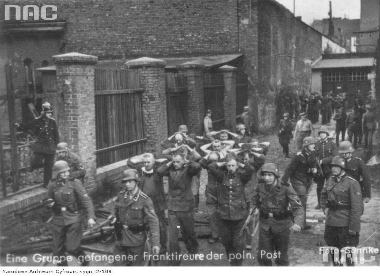 Een zwart-wit foto van Duitse Wehrmacht soldaten die een groep Poolse krijgsgevangenen afvoeren.