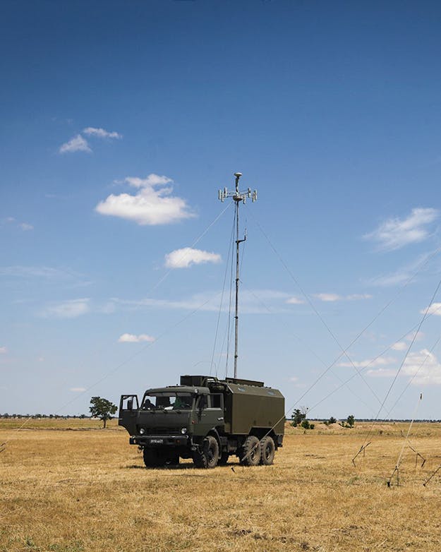 Het Russische voertuig R-330Zh Zhitel kan satellietnavigatie tot op honderden kilometers verstoren. Een dor landschap met een militair voertuig en een bouwwerk met grote antennes.