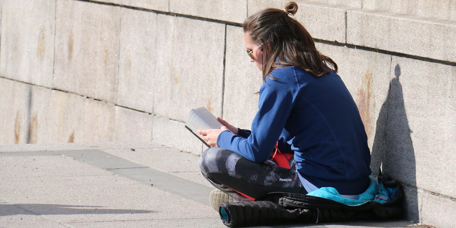 Een persoon zit op een jas op de grond in kleermakerszit een boek te lezen.