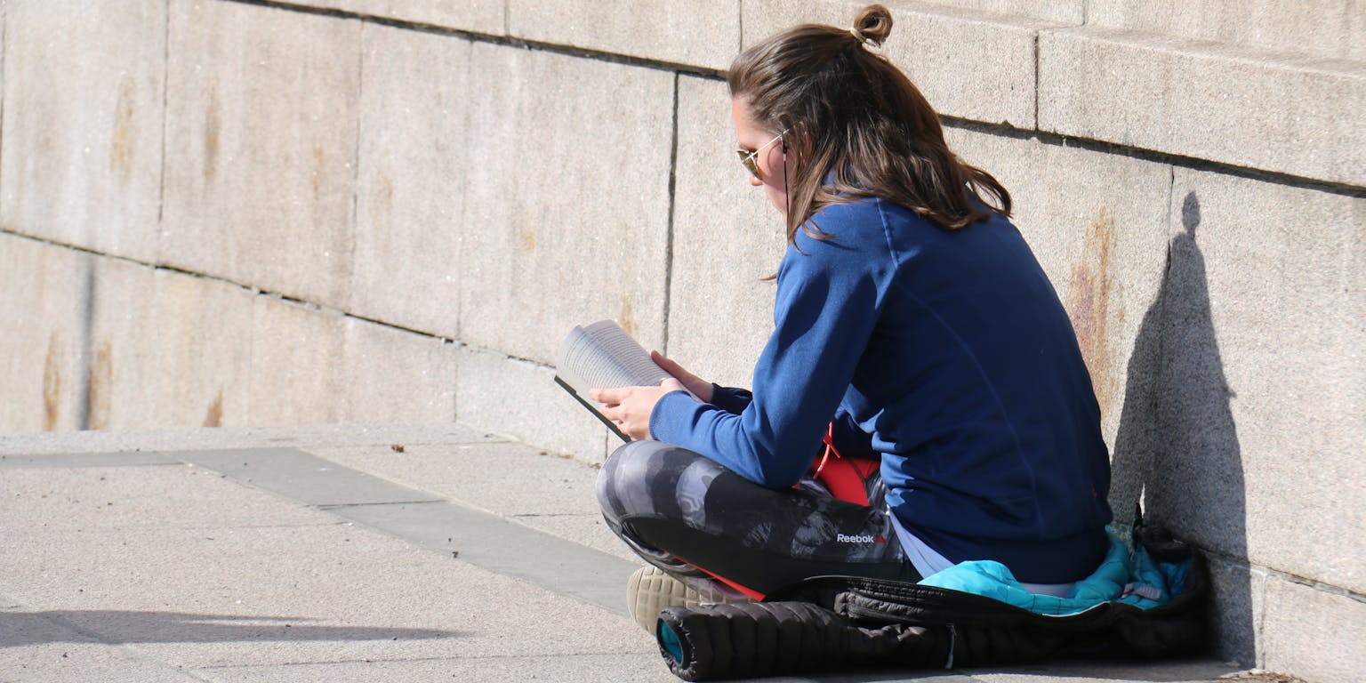 Een persoon zit op een jas op de grond in kleermakerszit een boek te lezen.