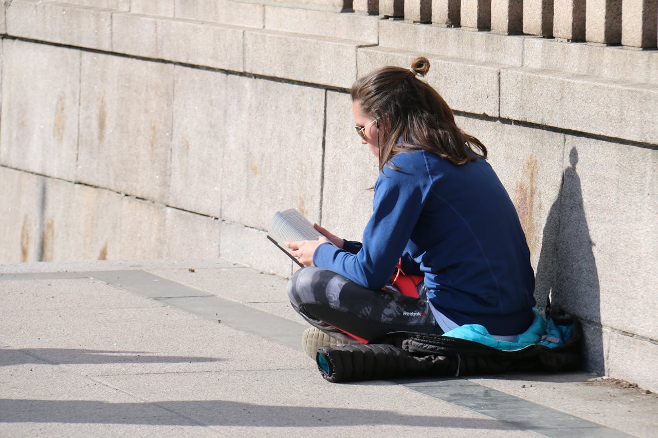 Een persoon zit op een jas op de grond in kleermakerszit een boek te lezen.