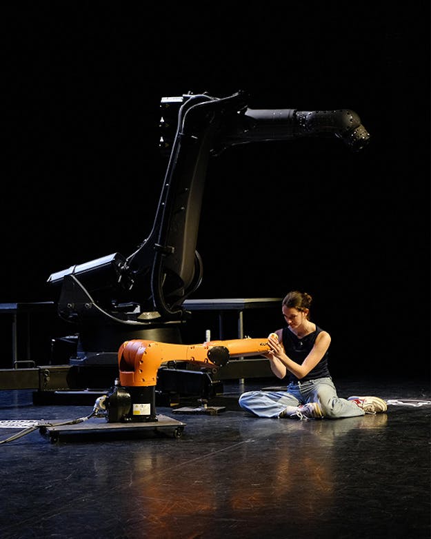 Rrepetitiemoment uit ‘Robot van de Rommelmarkt’ van Bram Ellen. Een donker toneelpodium met een vrouw die op de grond zit naast een oranje robotarm.
