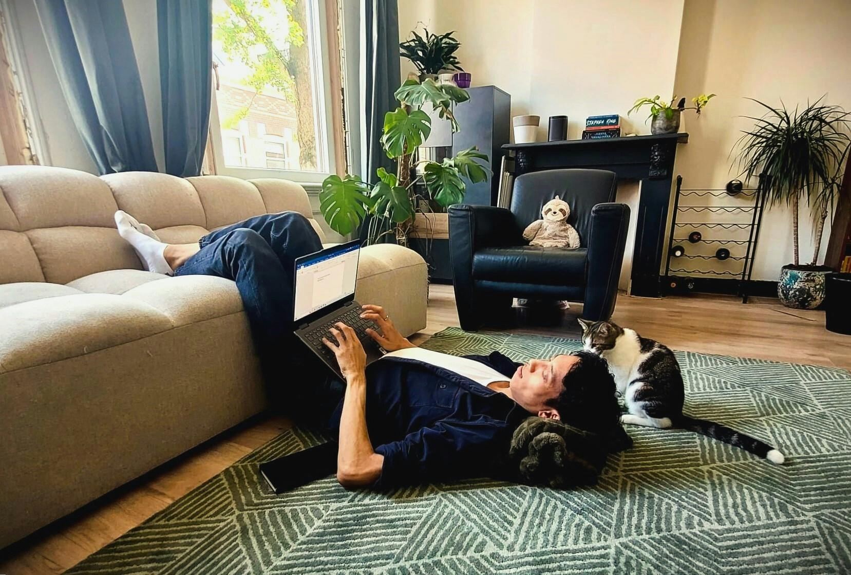 Face of Science Patrick Tang schrijft ondersteboven hangend op zijn bank een wetenschappelijk artikel op zijn laptop, terwijl zijn kat toekijkt