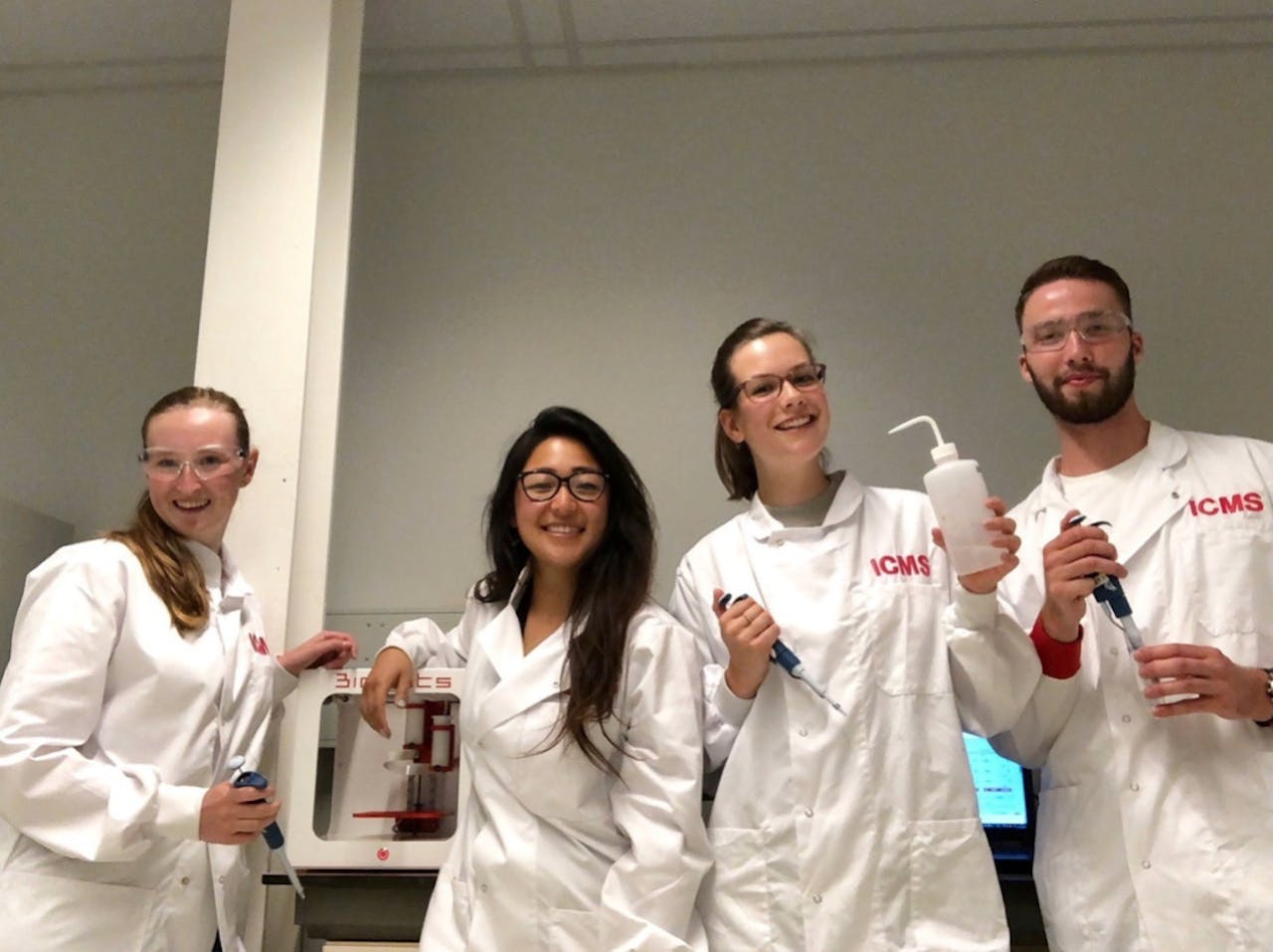 Master- en bachelor-studenten begeleiden hoort bij mijn werk. Hier ben ik een masterstudent aan het begeleiden in haar onderzoek en twee bachelorstudenten aan het leren hoe zij zelfstandig onderzoek moeten doen. Vier studenten met een witte labjas aan.