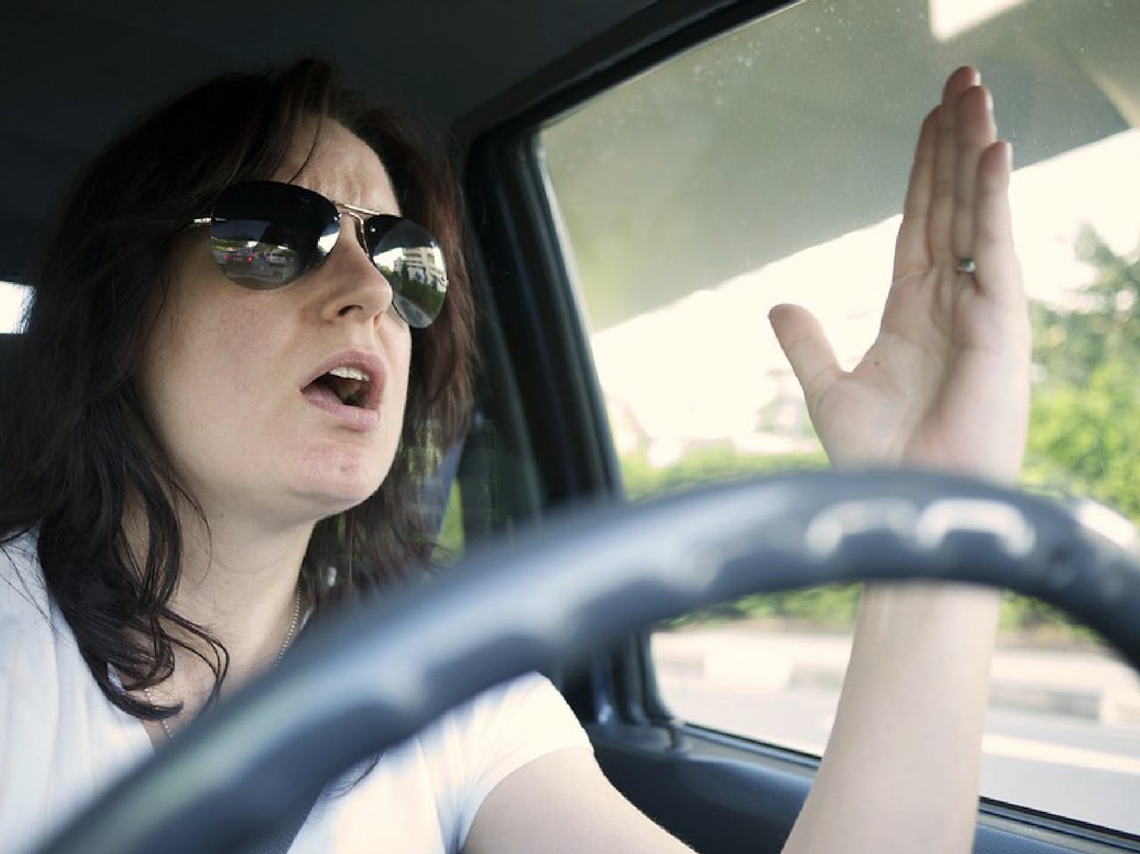 Een vrouw achter het stuur van een auto steekt haar hand op.