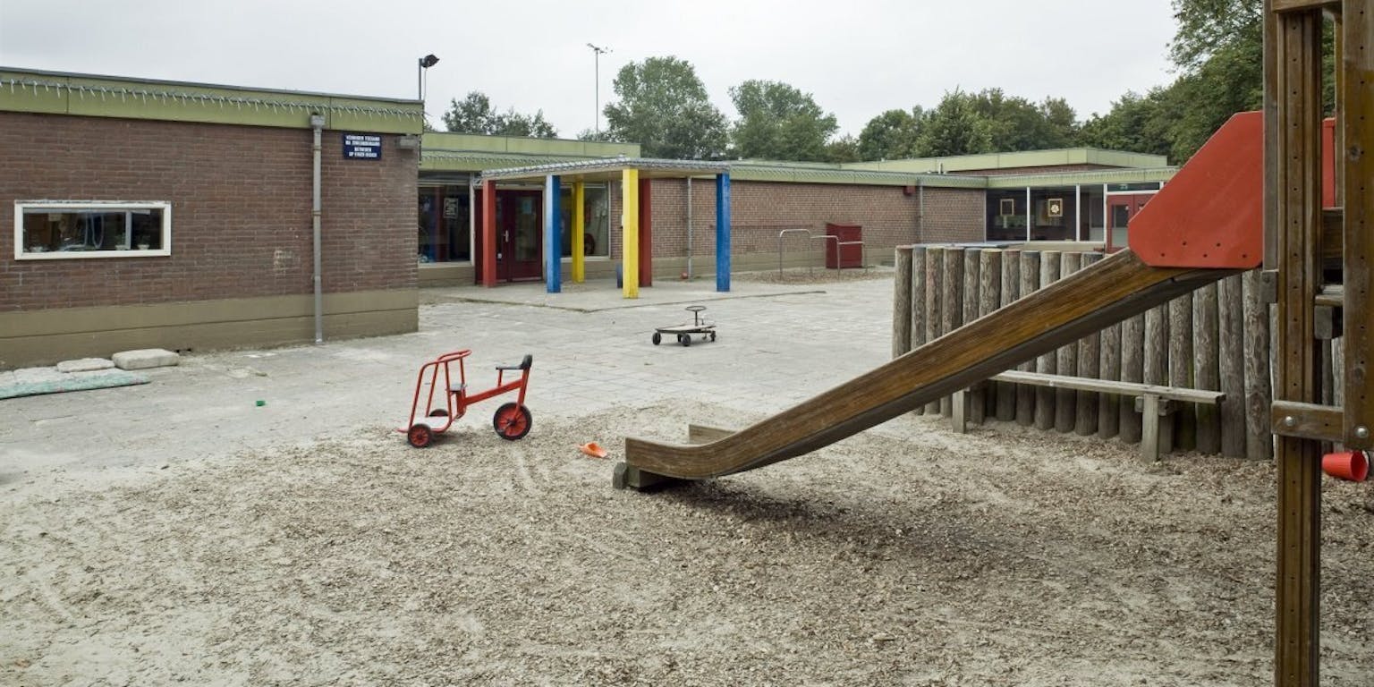 Een leeg schoolplein met een glijbaan en trapfiets met een school op de achtergrond.