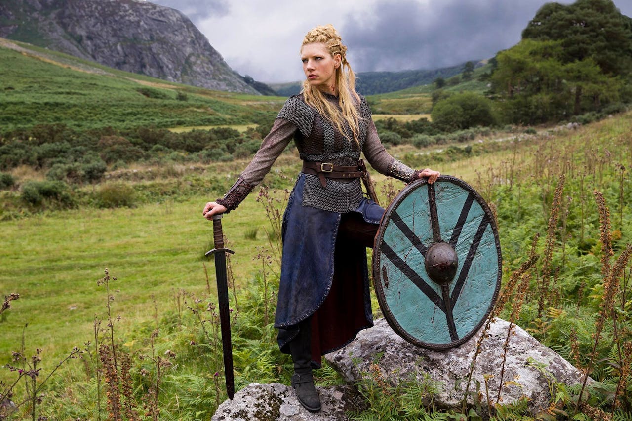 Een vrouw in een vikingkostuum staat in een veld met een schild en een zwaard.