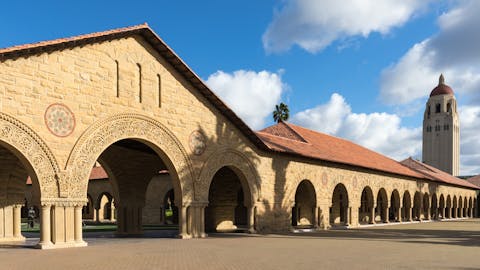 Standford University ligt in het hart van Silicon Valley. Een zandkleurig gebouw met bogen en een toren, tegen een blauwe lucht met een paar witte wolken.