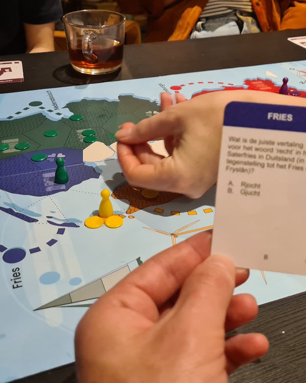 Het bordspel ‘Streektaalstrijd’ van de Rijksuniversiteit Groningen. Mensen spelen het bordspel Streektaalstrijd. Op de voorgrond een handje die één van de vraagkaartjes vasthoudt.