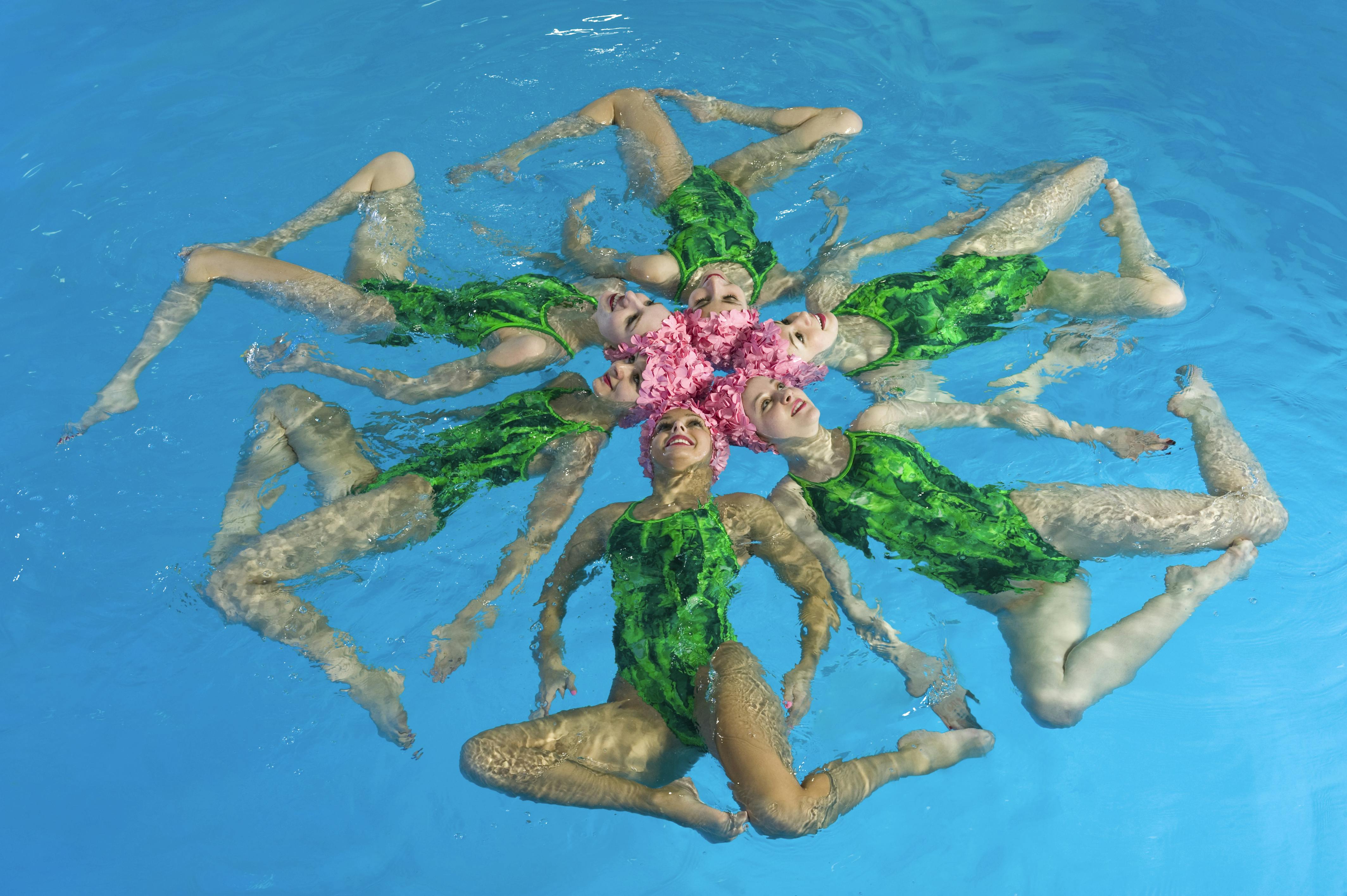 Een groep vrouwen in een zwembad met een groen badpak en bloemen op het hoofd zwemmen synchroon.