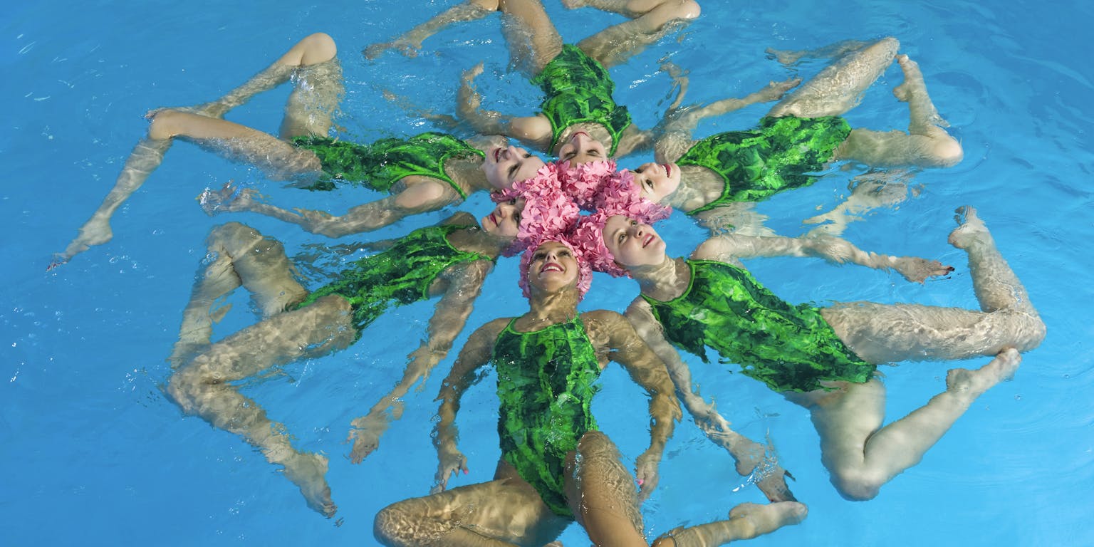 Een groep vrouwen in een zwembad met een groen badpak en bloemen op het hoofd zwemmen synchroon.