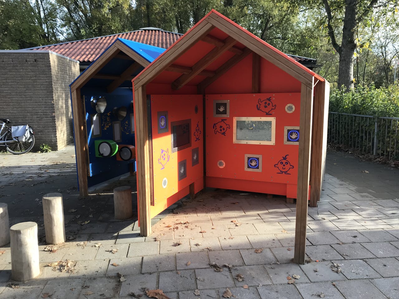 Twee houten speelhuisjes in een park met een blauwe en oranje dak.