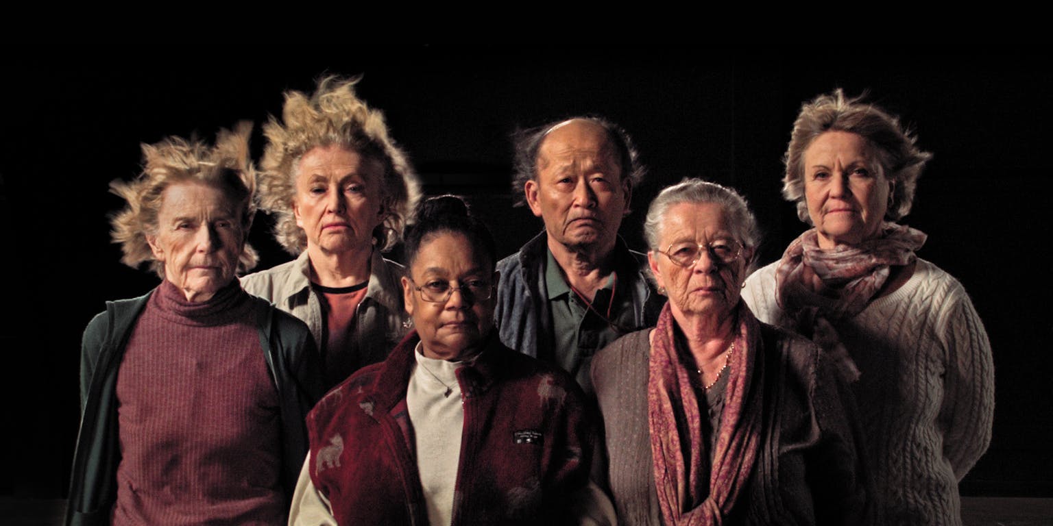 Zes personen op een rij kijken in de camera. De achtergrond is zwart.