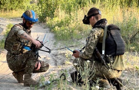 Oekraïense soldaten testen hun geïmproviseerde aanvaldrones uit. Twee militairen zitten gehurkt op een zandpad. Ze hebben beiden een drone in hun handen.