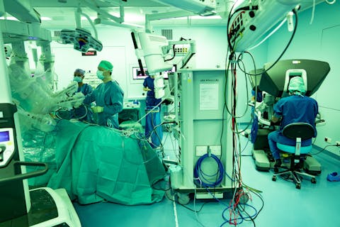 Robotchirurgie ziet er anders uit dan we gewend zijn van tv. De chirurg staat niet aan de operatietafel, maar zit helemaal rechts, in een apart controlekamertje, waar hij de operatierobot bedient die boven de patiënt hangt. Foto van een OK. Er staan twee operatieassistenten bij de patiënt. Boven de patiënt hangen vier robotarmen. Helemaal rechts zit een chirurg achter het controlepaneel van de operatierobot.