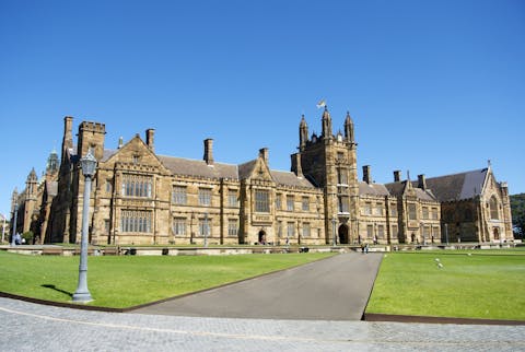 Een van de historische gebouwen op de campus van de University of Sydney. Een groot klassiek gebouw met torentjes. Het uitgestrekt grasveld dat ervoor ligt, wordt doorsneden door een asfaltweg. De lucht is strakblauw.